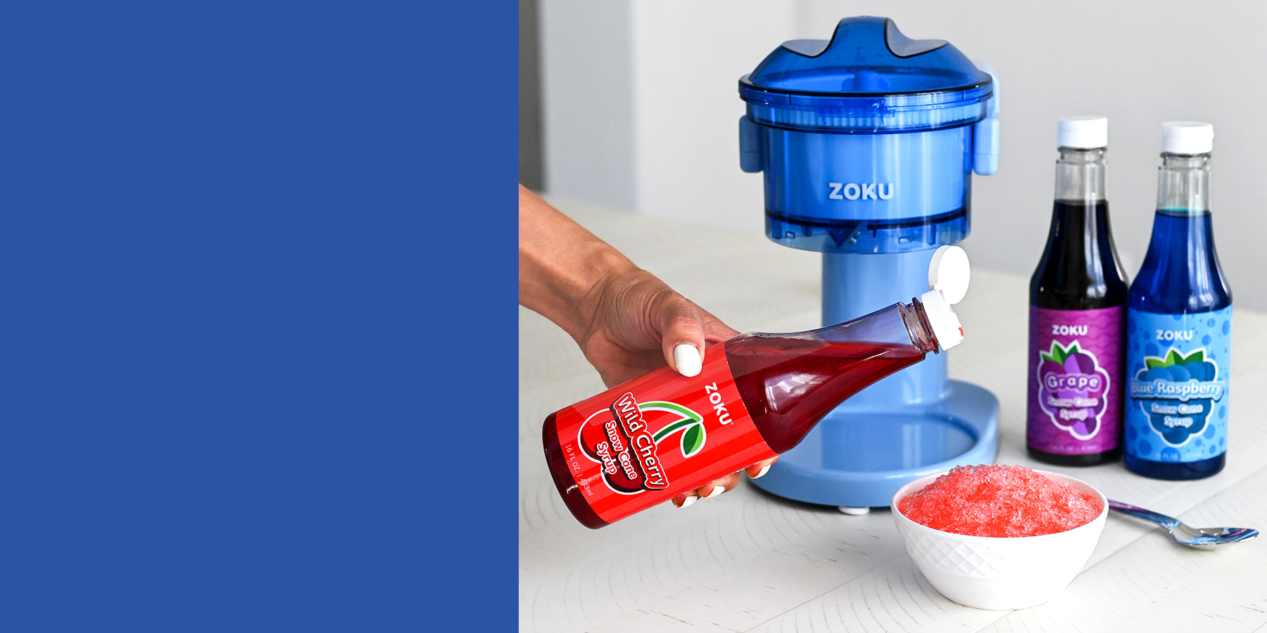 Hand pouring ZOKU Wild Cherry Snow Cone Syrup into a bowl of shaved ice with a blue ZOKU ice shaver machine and bottles of ZOKU Grape and Blue Raspberry Snow Cone Syrups in the background.